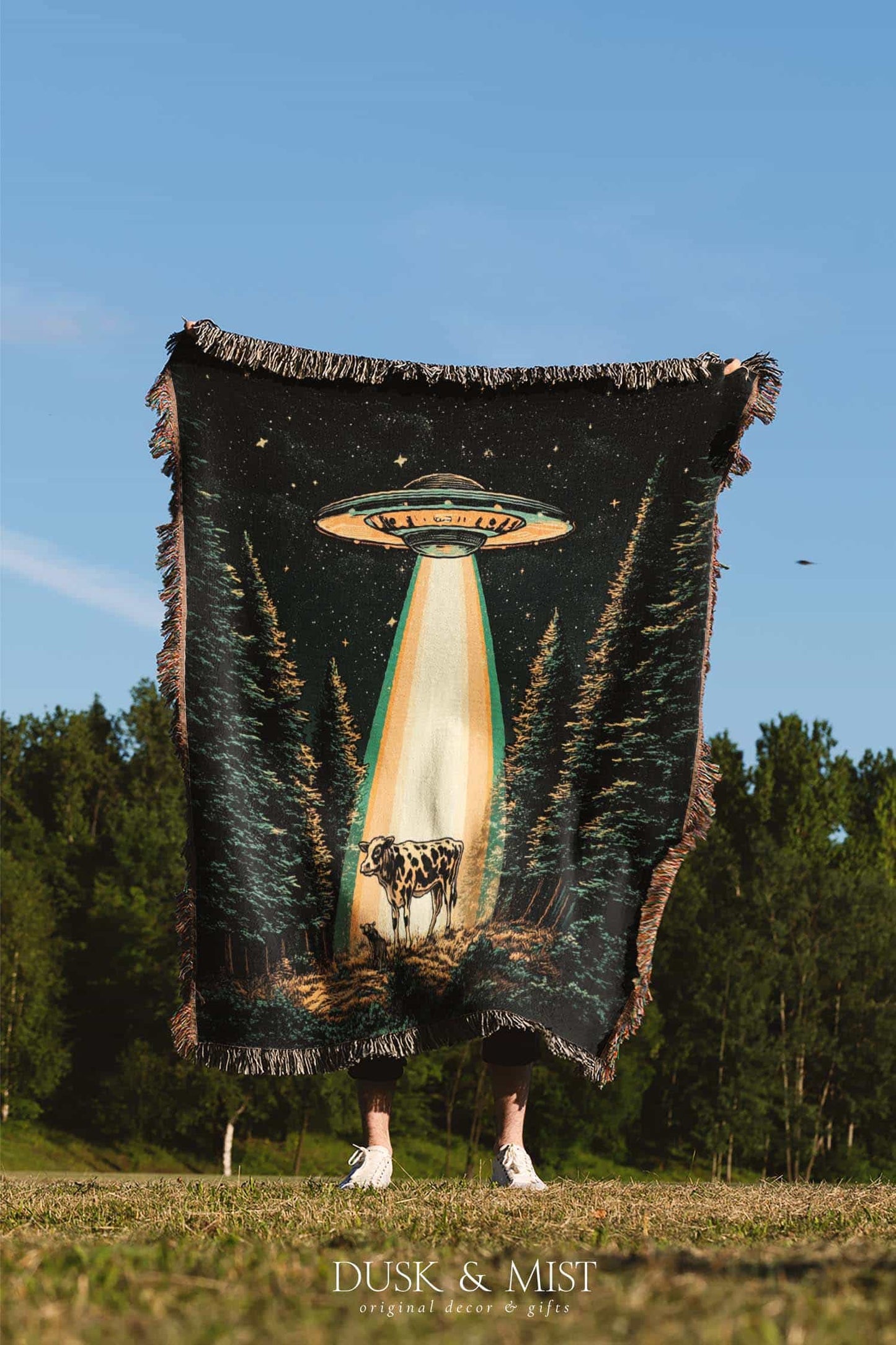 Woven cotton blanket showing a UFO shining a beam of light onto a cow in a forest clearing under a starry night sky.
