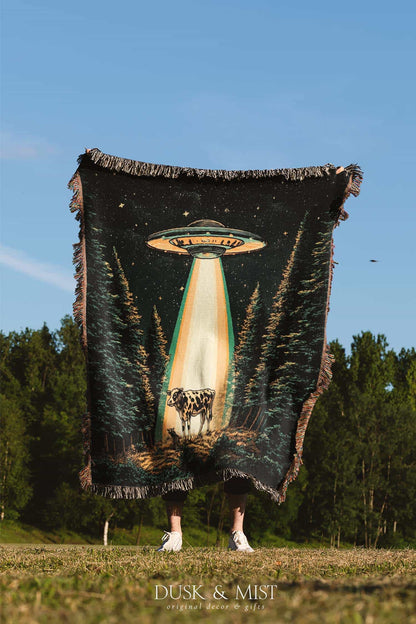 Woven cotton blanket showing a UFO shining a beam of light onto a cow in a forest clearing under a starry night sky.