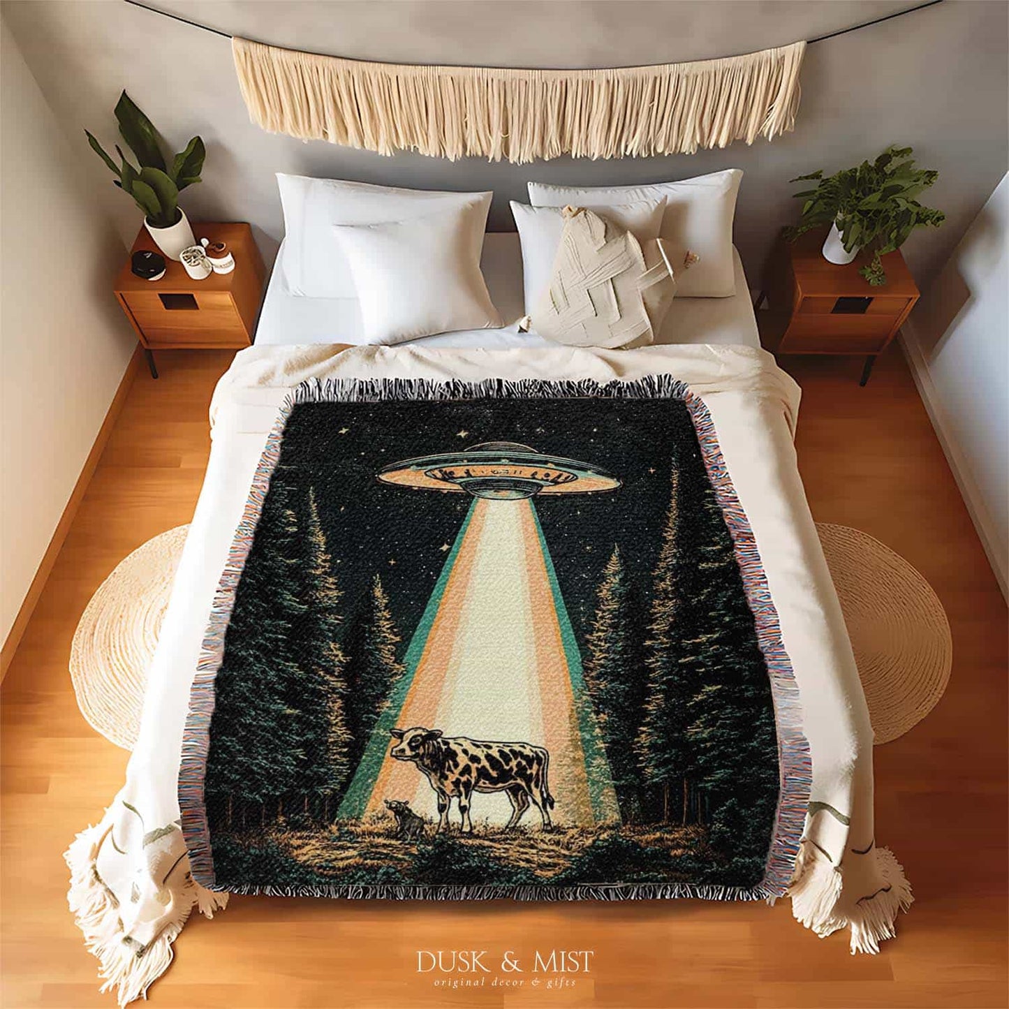 Woven cotton blanket showing a UFO shining a beam of light onto a cow in a forest clearing under a starry night sky.