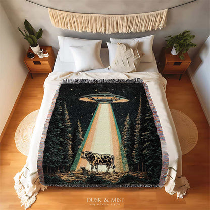 Woven cotton blanket showing a UFO shining a beam of light onto a cow in a forest clearing under a starry night sky.