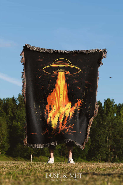 Woven cotton blanket featuring a UFO with a fiery orange beam blasting into a dark forest, with glowing explosions and cosmic night details.