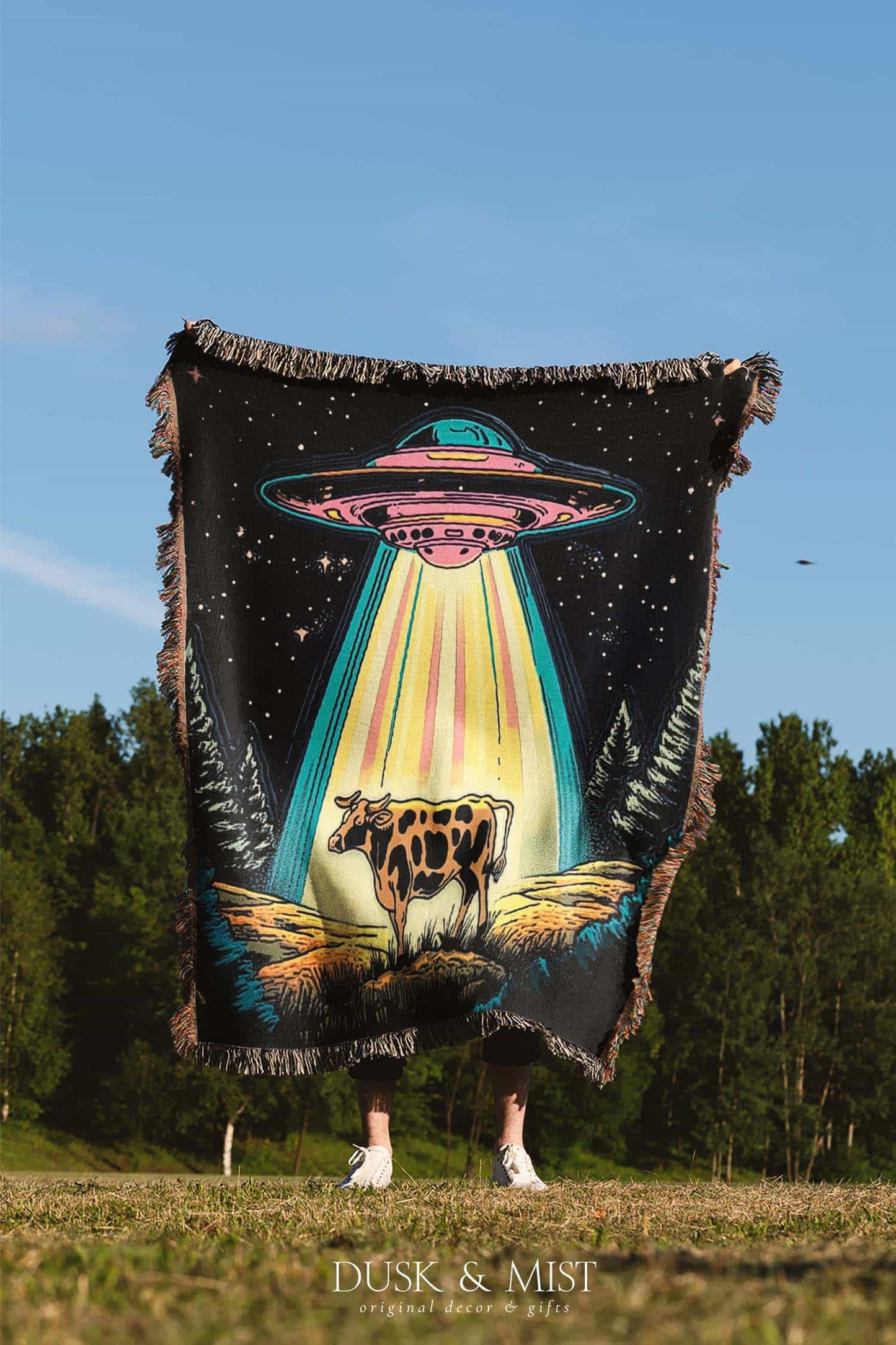 Blanket with a UFO abduction of a cow design against a starry night sky.