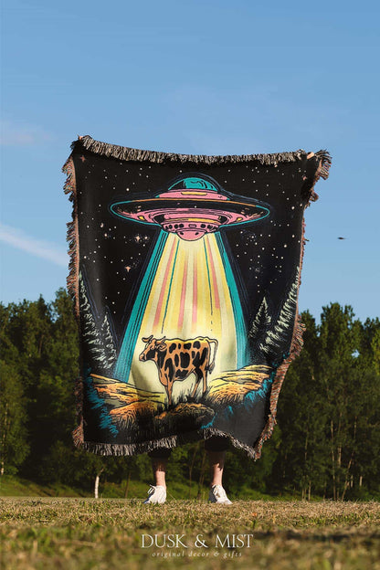 Blanket with a UFO abduction of a cow design against a starry night sky.